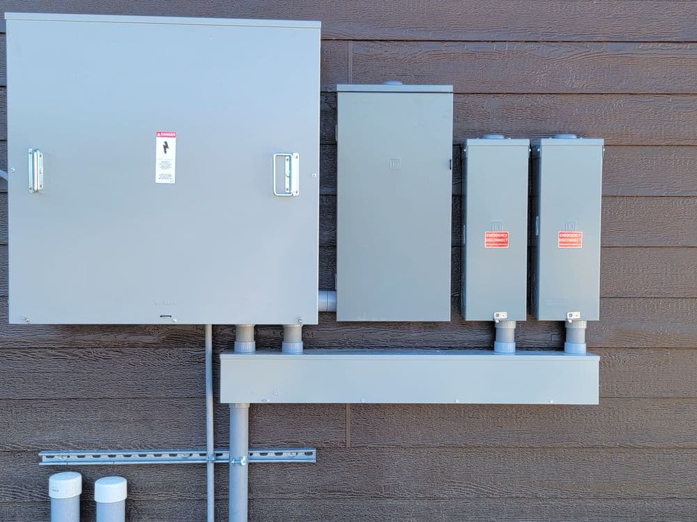 Electrical panels mounted on a wooden wall with conduit pipes and safety labels.
