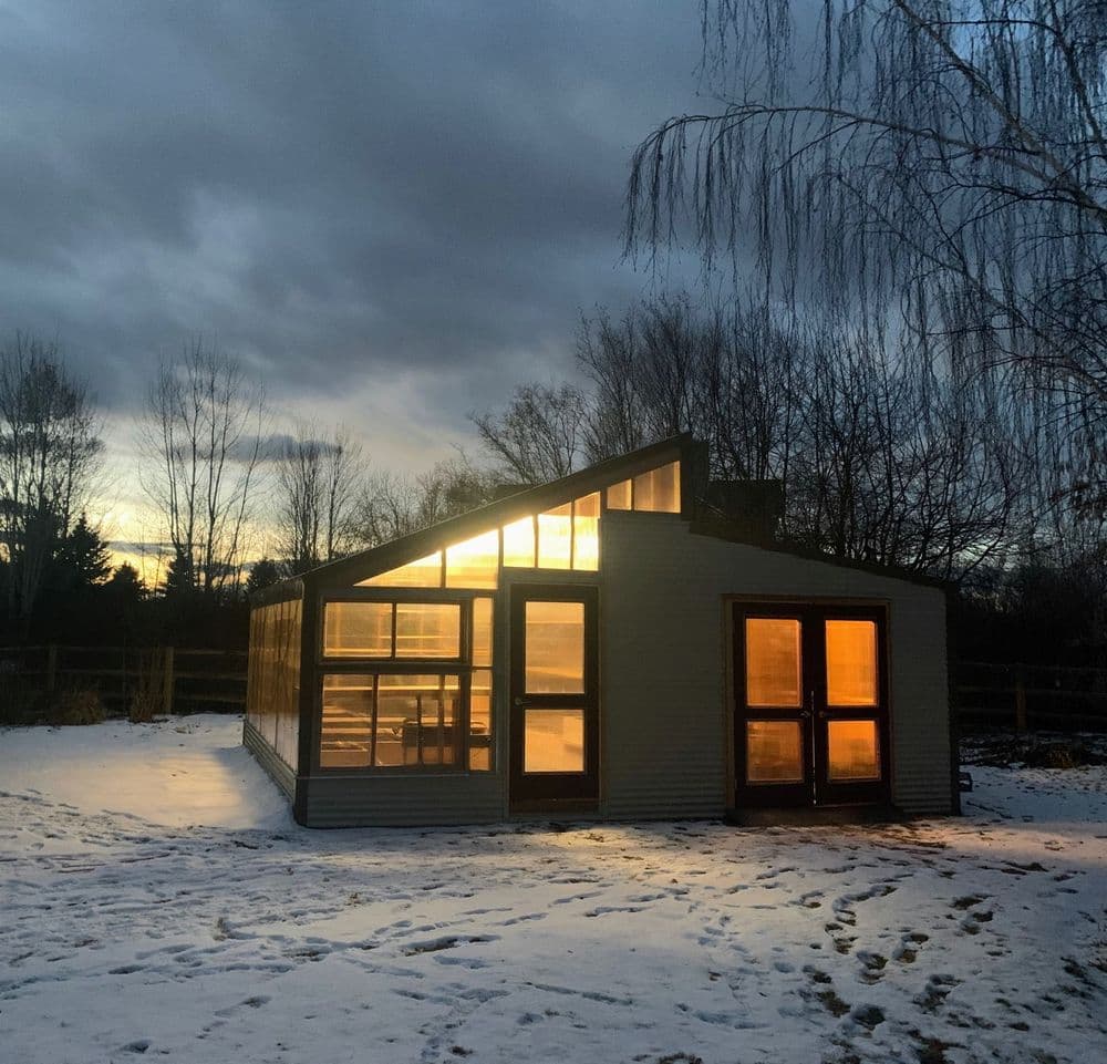 Modern greenhouse illuminated at sunset amidst a snowy landscape with bare trees.