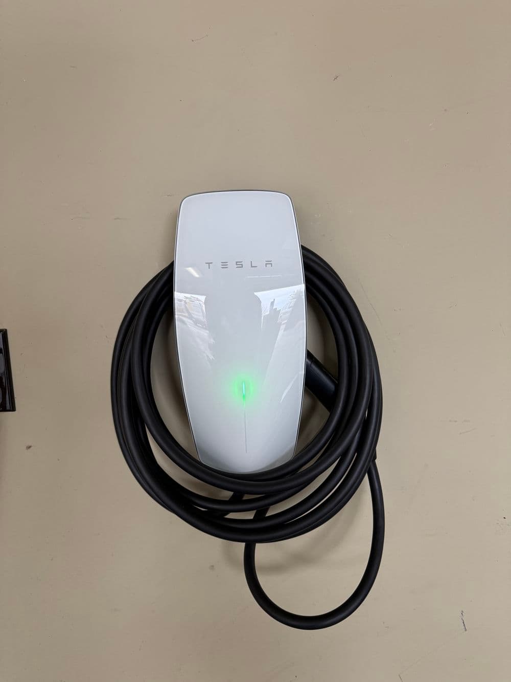Tesla electric vehicle charger with green indicator light and coiled cable on a neutral background.
