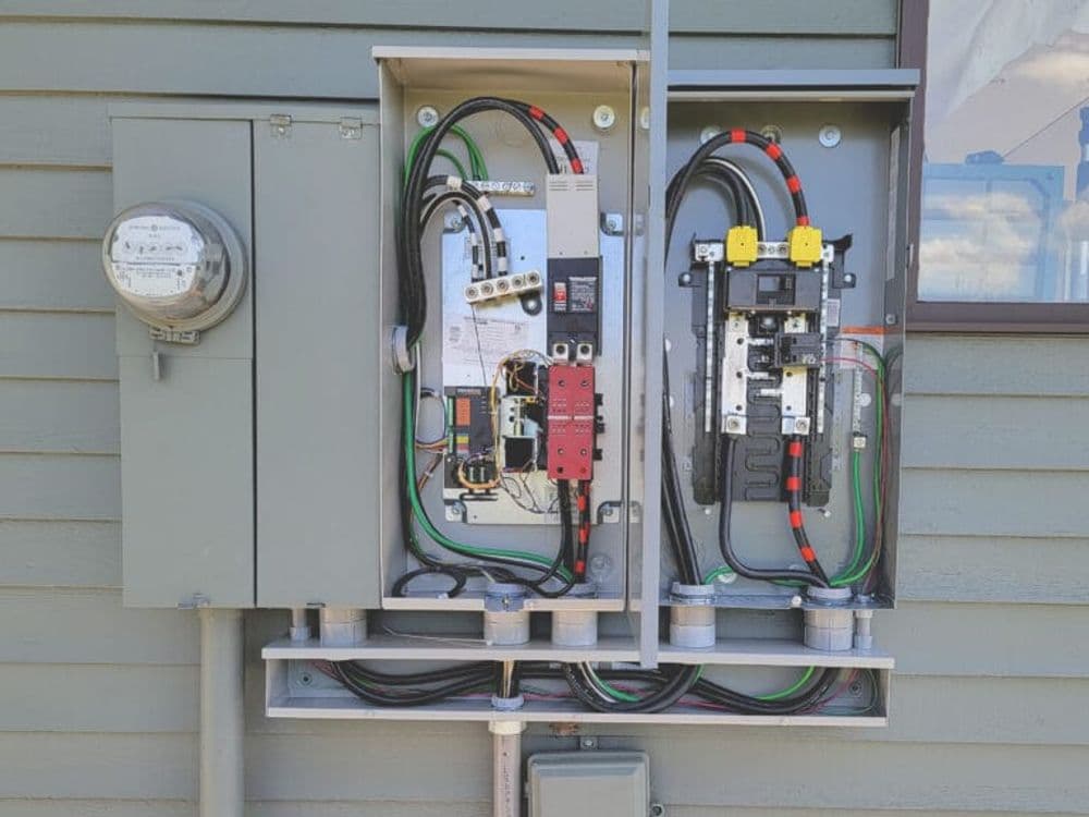 Electrical panel installation with circuit breakers and meter on a house exterior wall.
