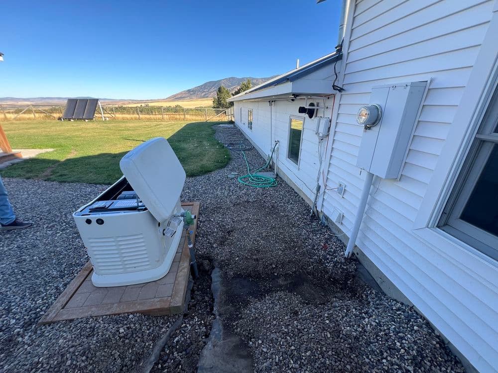 Generator installation beside a white house, with mountains and solar panels in the background.