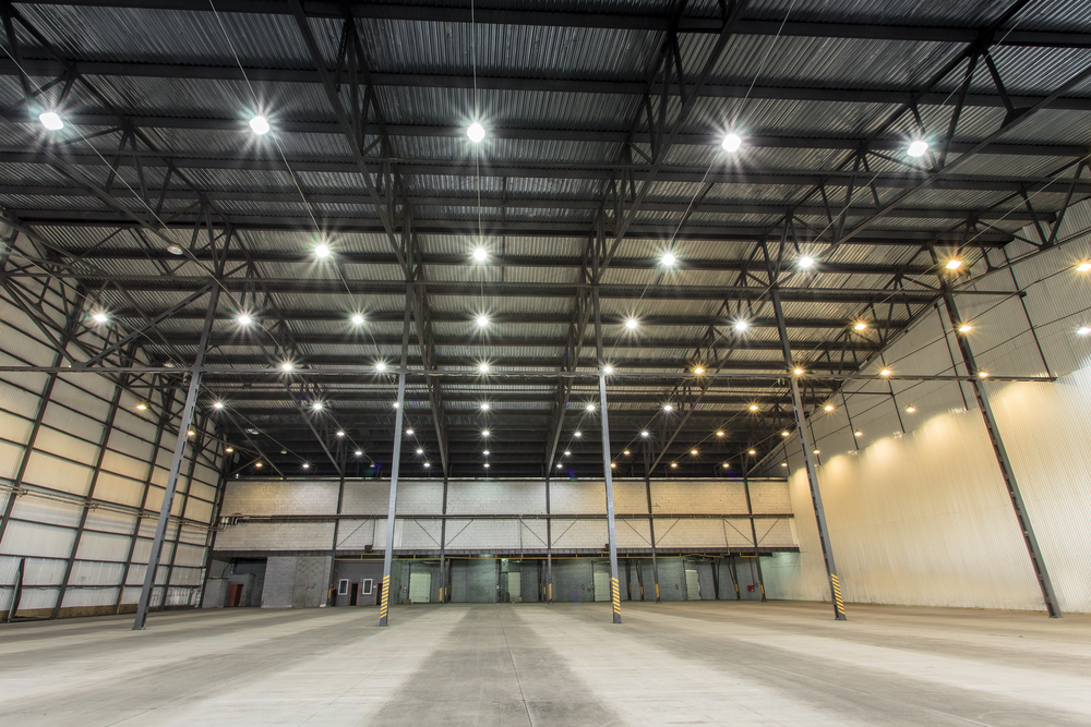 Wide view of a large, empty warehouse with high ceilings, metal beams, and bright overhead lights expertly installed by an electrician, plus multiple loading dock doors along the back wall.