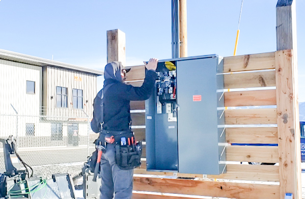 An electrician wearing a tool belt works on an open electrical panel mounted on a wooden wall outdoors near an industrial building.