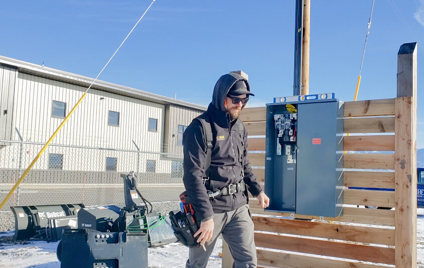 An electrician in outdoor winter clothing stands near an open electrical panel mounted on a wooden fence, with industrial buildings and equipment in the background.