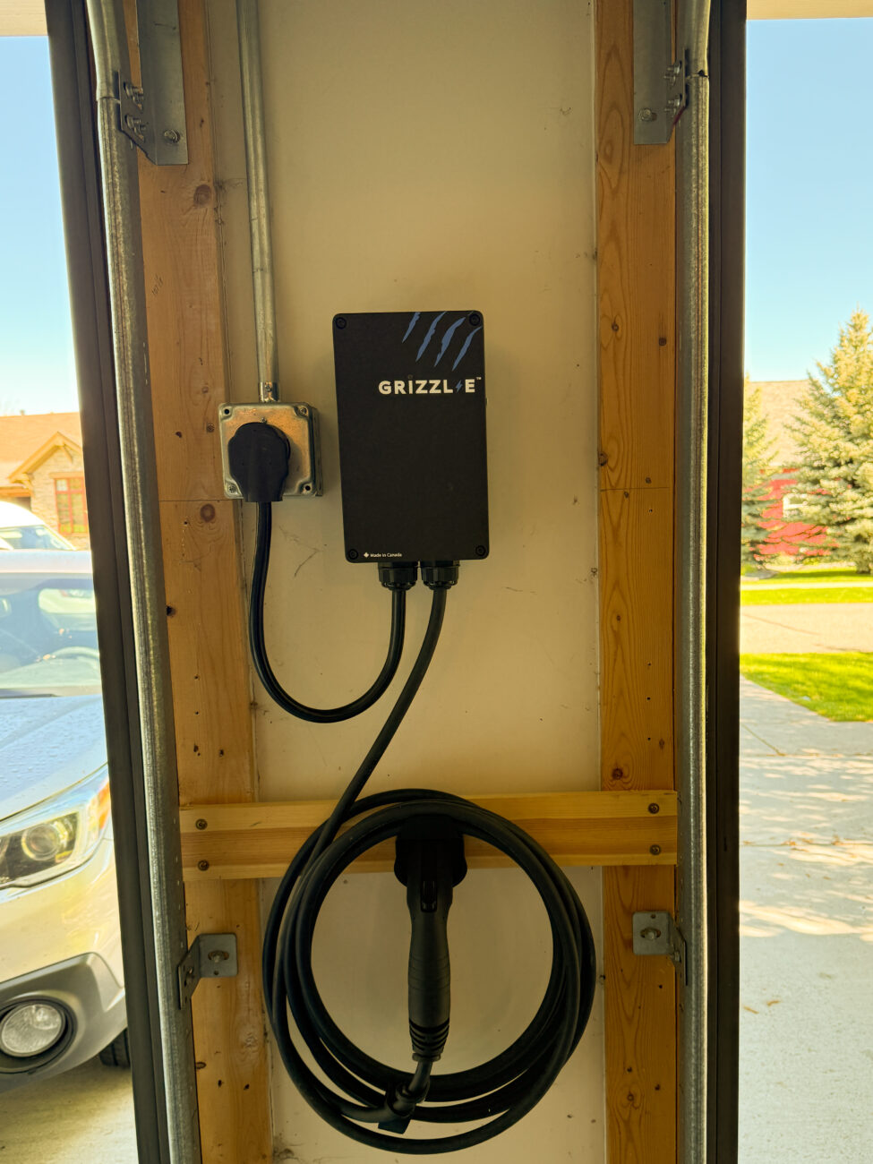 A Grizzl-E electric vehicle charging station, professionally installed by an electrician, is mounted on a garage wall, with its charging cable neatly coiled on a wooden holder below.