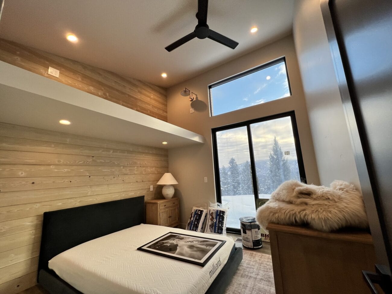 A modern bedroom with wood-paneled walls, a large window showing snowy trees, a bed with a framed photo, and unpacked items on the floor and dresser.