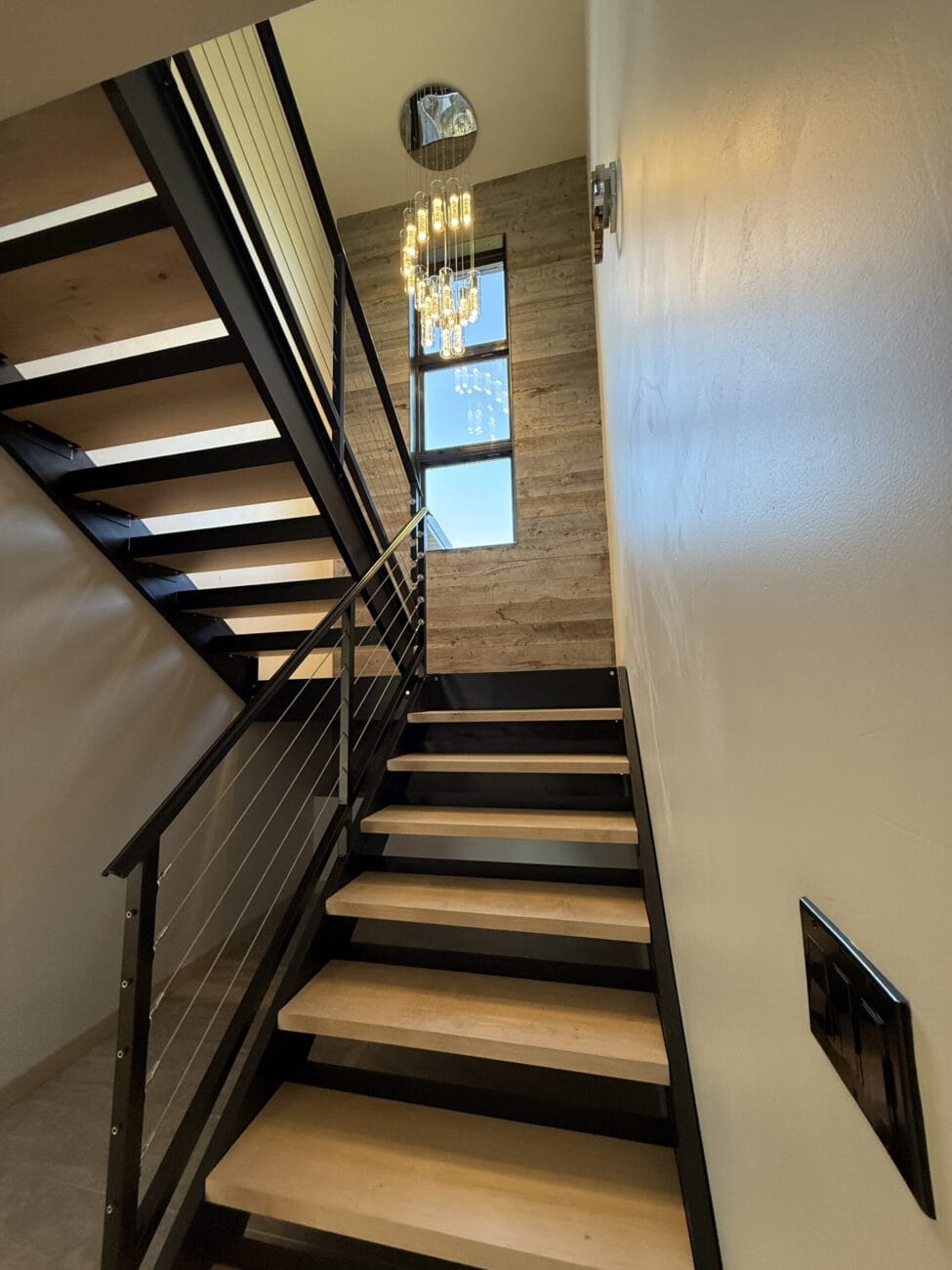 A modern staircase with light wood steps, black metal railing, cable balusters, and a multi-tiered chandelier hanging by large vertical windows.