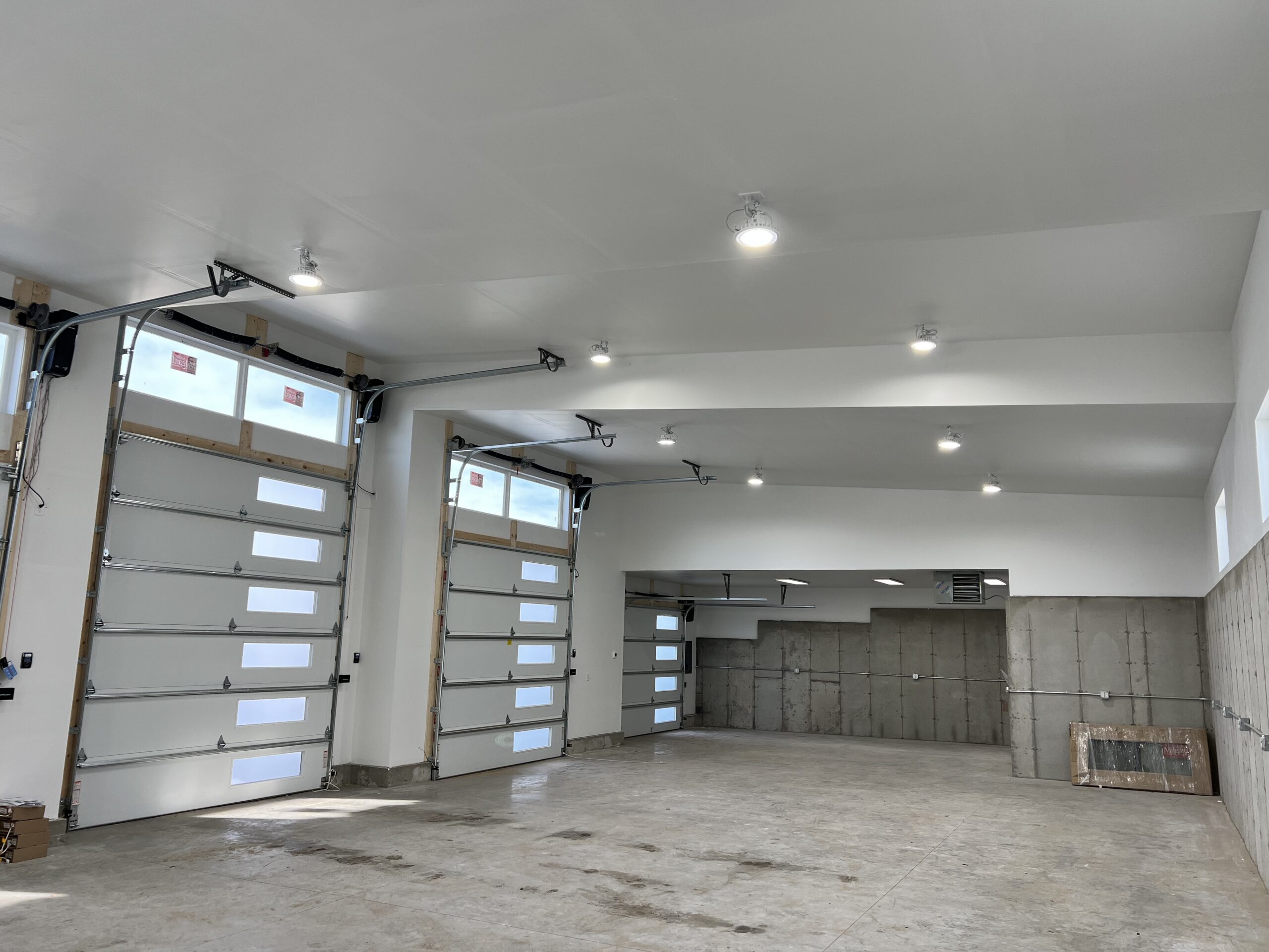 A spacious, empty garage with white walls, concrete floors, multiple ceiling lights installed by a professional electrician, and three large glass-paneled overhead doors.
