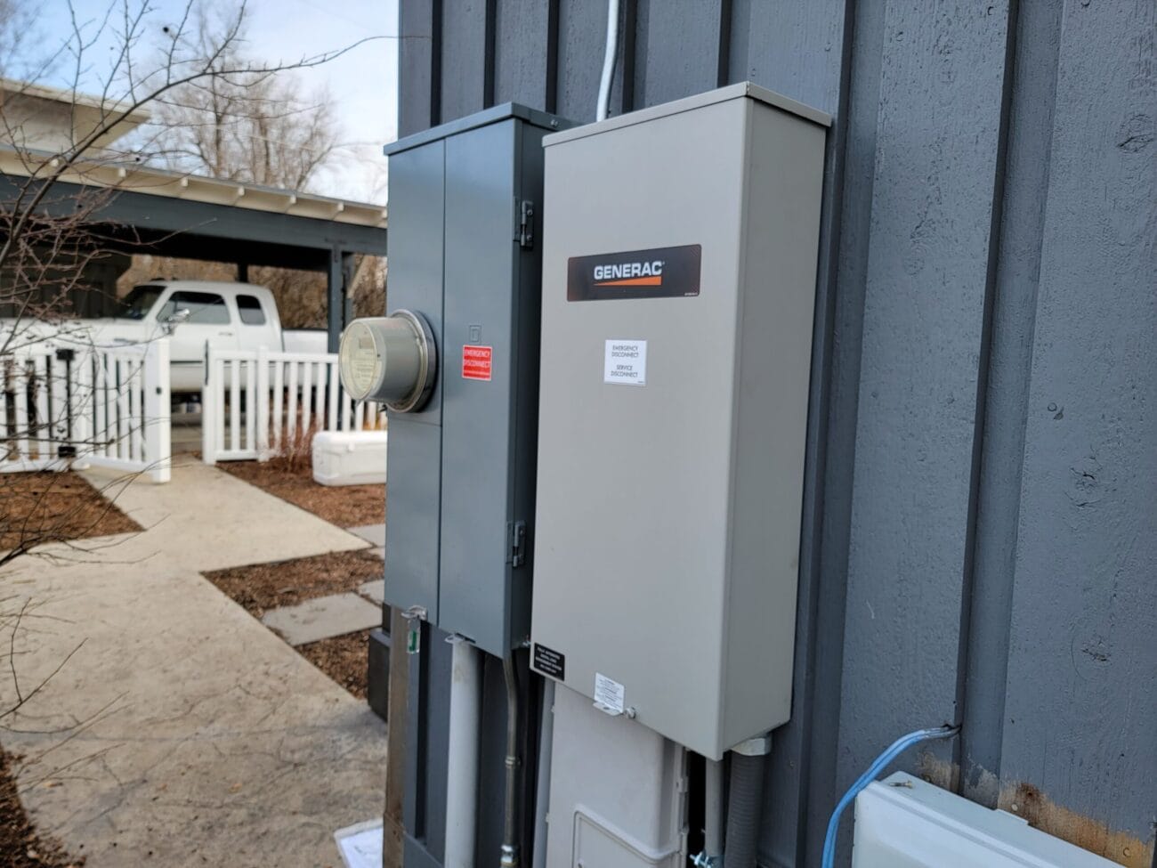 A Generac electrical panel and a utility meter are mounted on the exterior wall of a building, next to a walkway and a white fence.