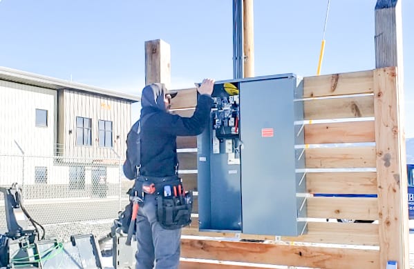 An electrician wearing a tool belt works on an open electrical panel mounted on a wooden fence outdoors, near a commercial building.