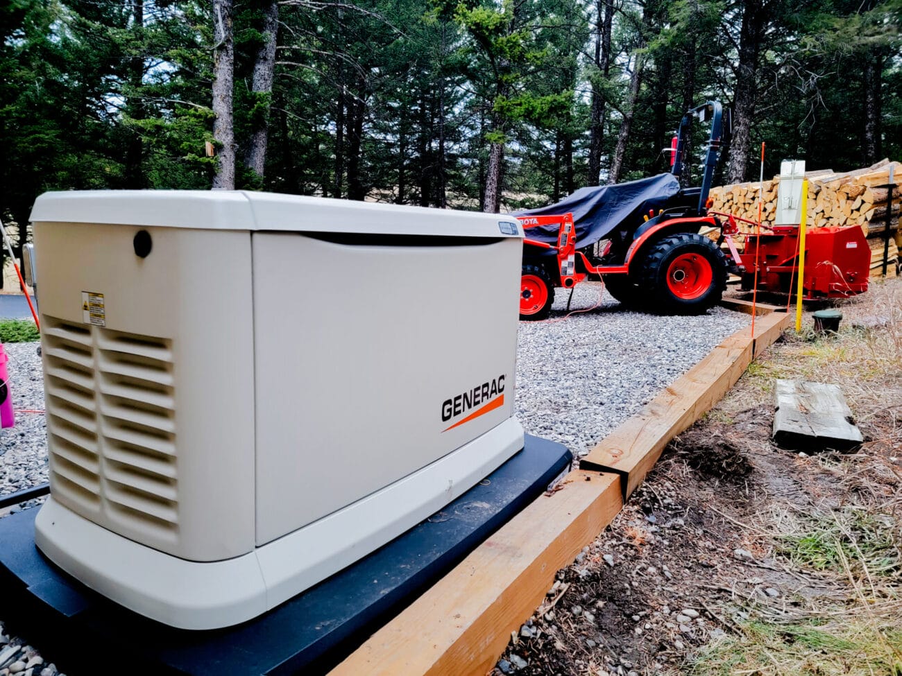 A Generac generator, installed by a skilled electrician, sits on a gravel pad near a wooded area, with a tractor and stacked firewood visible in the background.