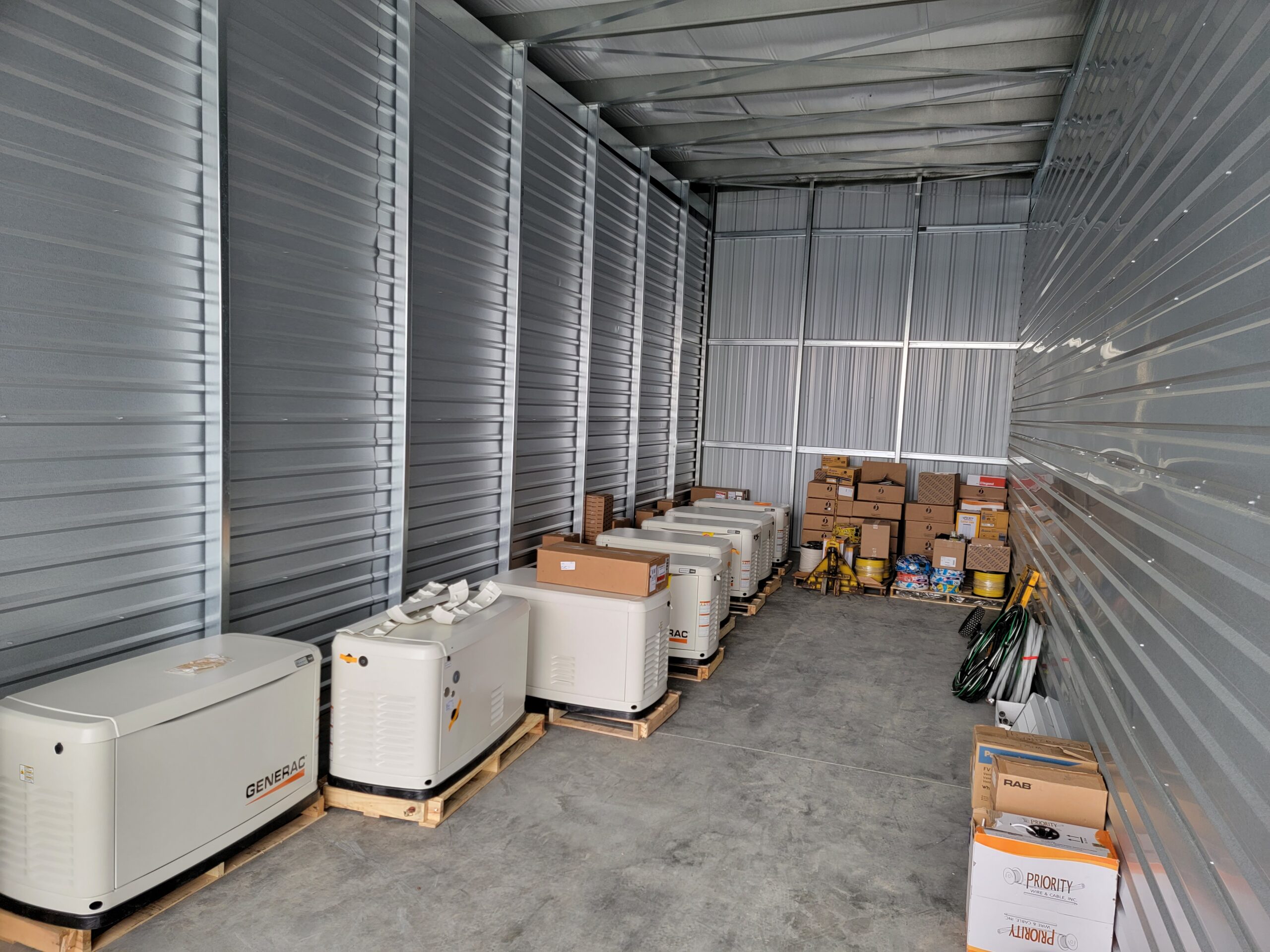 Several white generators are lined up inside a metal storage unit, with boxes, hoses, and other electrician supplies stacked along the back wall.