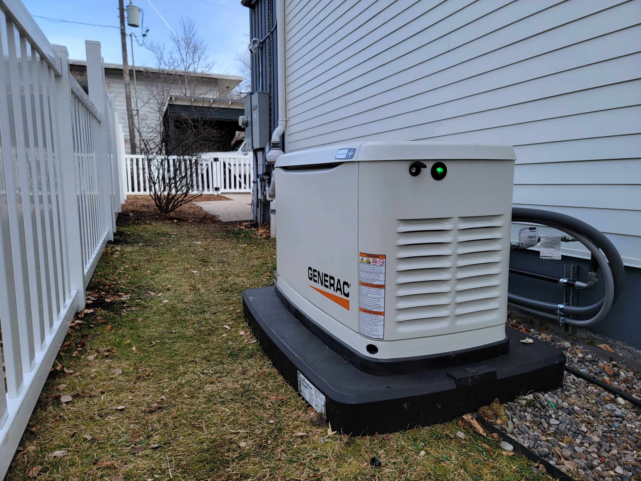 A Generac standby generator, professionally installed by an electrician on a black platform beside a white house, with utility connections and a white fence nearby.