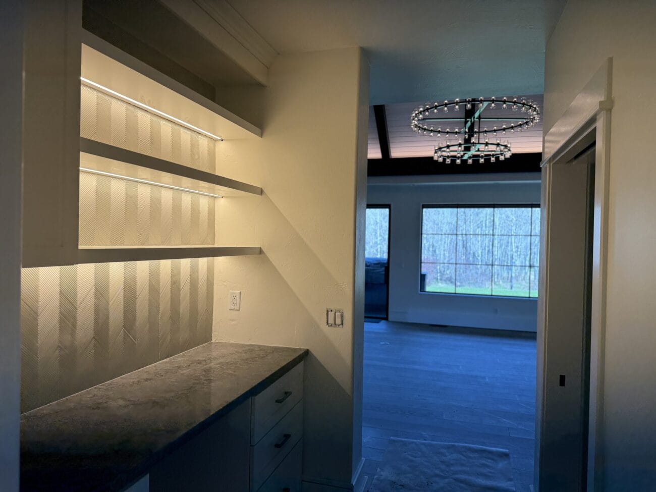 A hallway with built-in shelves and a countertop on the left, leading to a room with large windows and a modern circular chandelier.