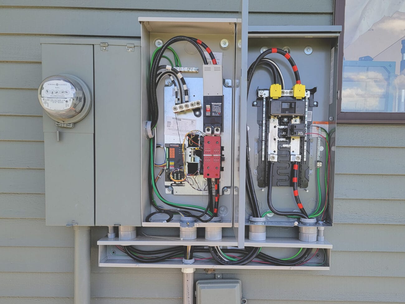 Two open electrical panels mounted on an exterior wall display circuit breakers, wiring, and meters—highlighting electrical service upgrades with color-coded cables and conduit connections clearly visible.