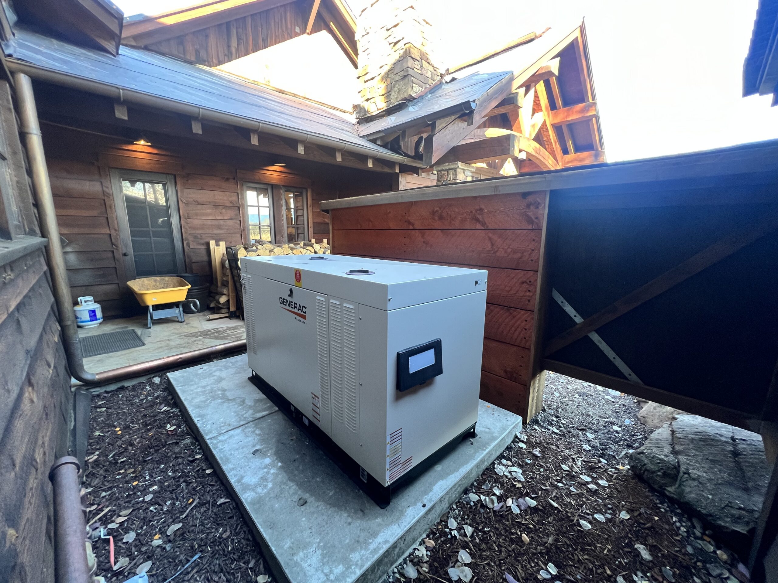 A large outdoor generator, installed by an electrician, sits on a concrete pad next to a wooden house with a wheelbarrow and various items nearby.