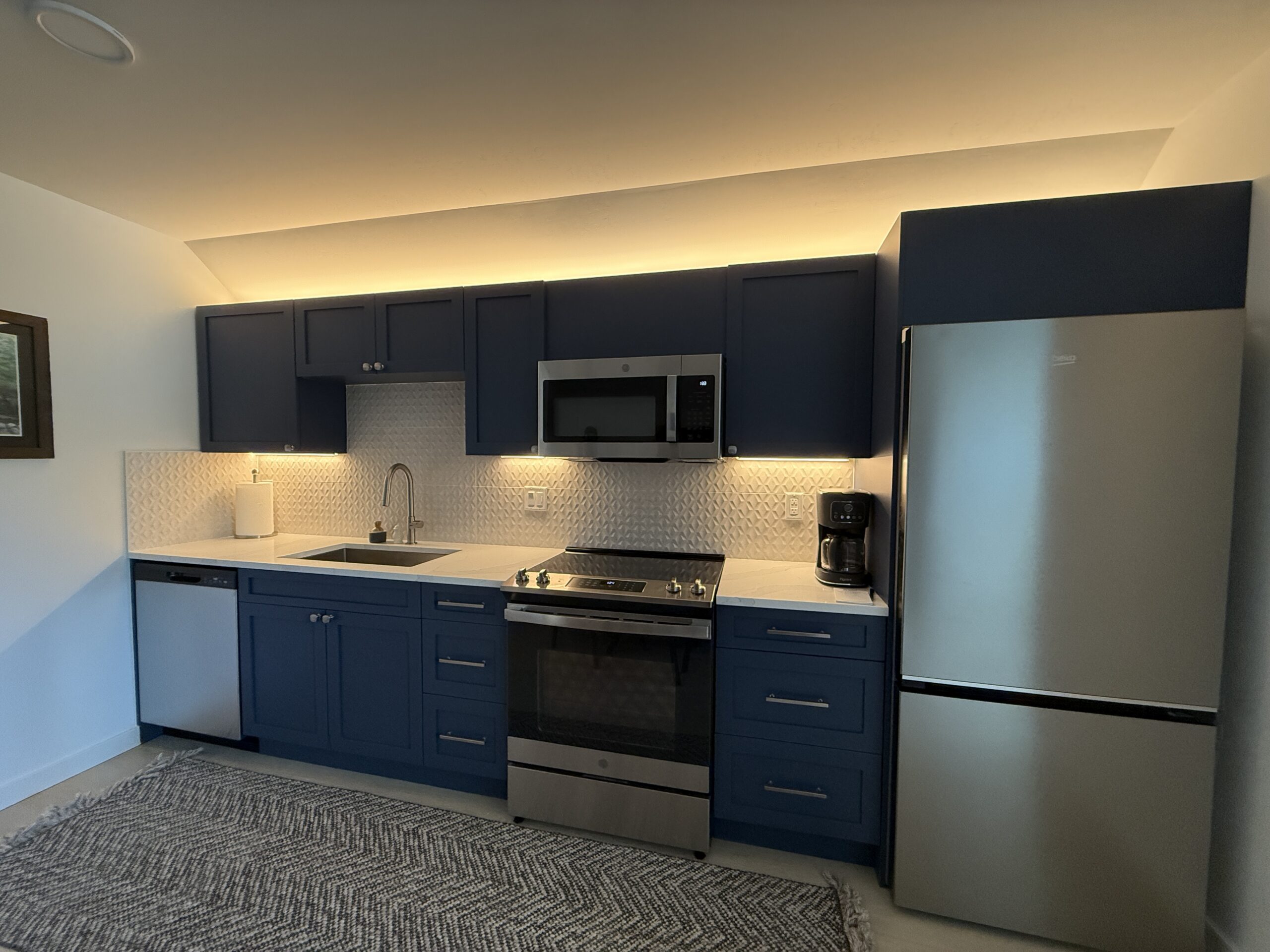 Modern kitchen with blue cabinets, stainless steel appliances, under-cabinet lighting installed by a skilled electrician, a sink, stove, microwave, dishwasher, refrigerator, and a patterned rug on the floor.