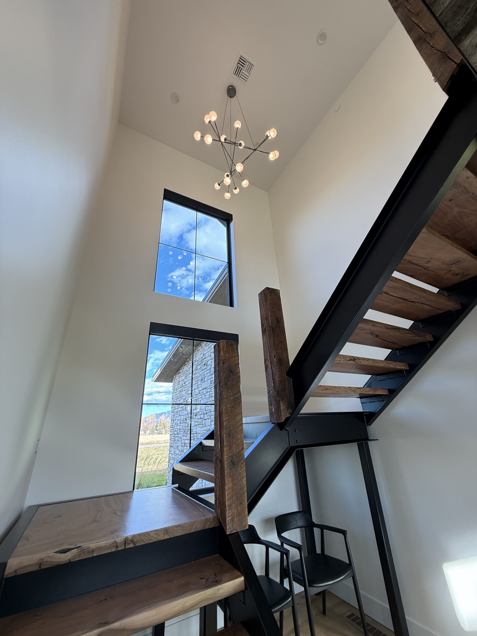 Modern staircase with wooden steps and black metal frame, large window with outdoor view, two black chairs, and a geometric chandelier overhead installed by a skilled electrician.