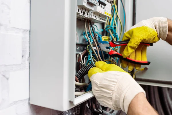 Person wearing yellow gloves uses pliers to work on electrical wires inside a circuit breaker panel during an electrical repair.