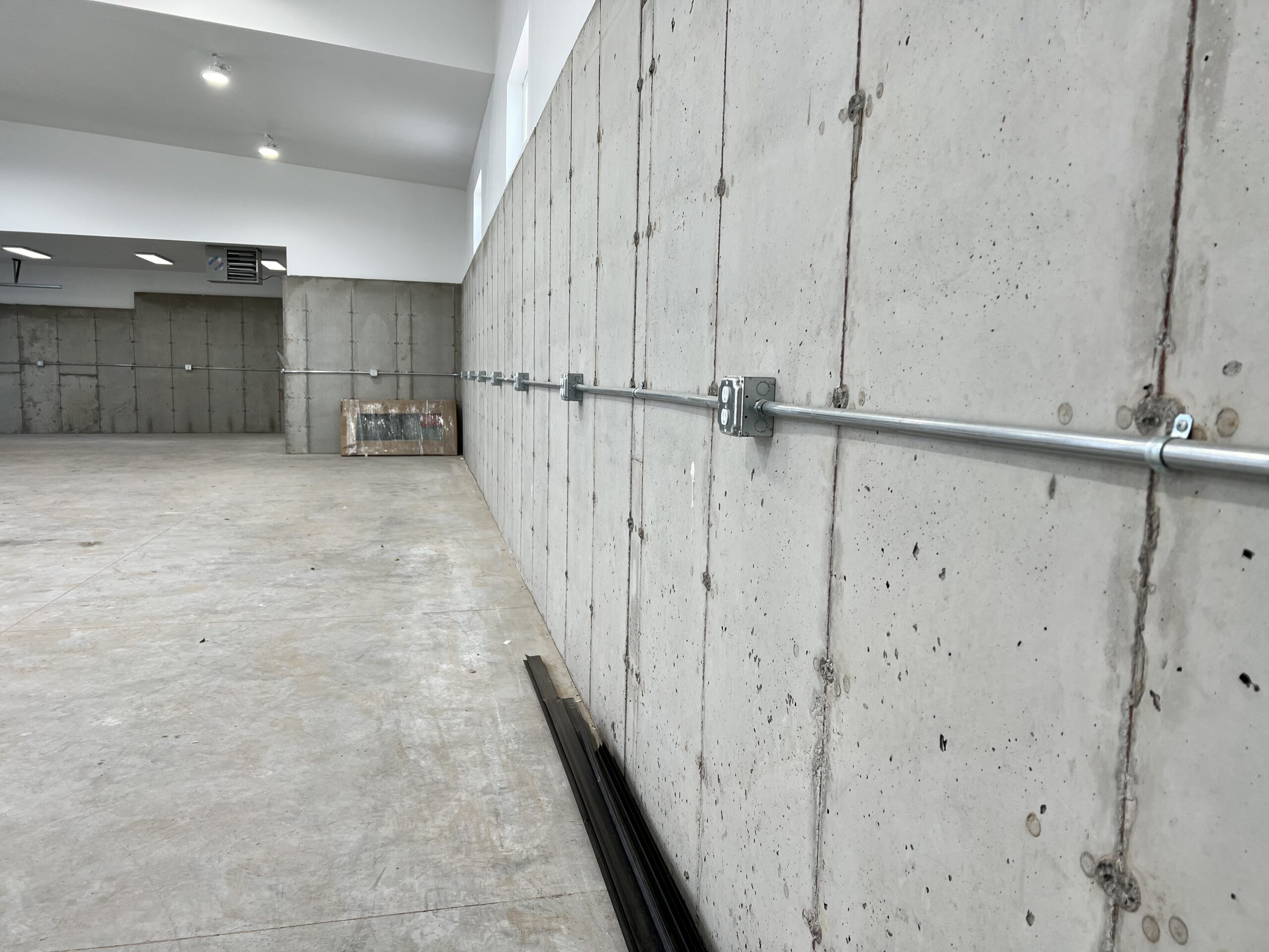 A large empty room with concrete walls and floor, exposed electrical conduit running along one wall—likely the work of an electrician—and construction materials in the background.