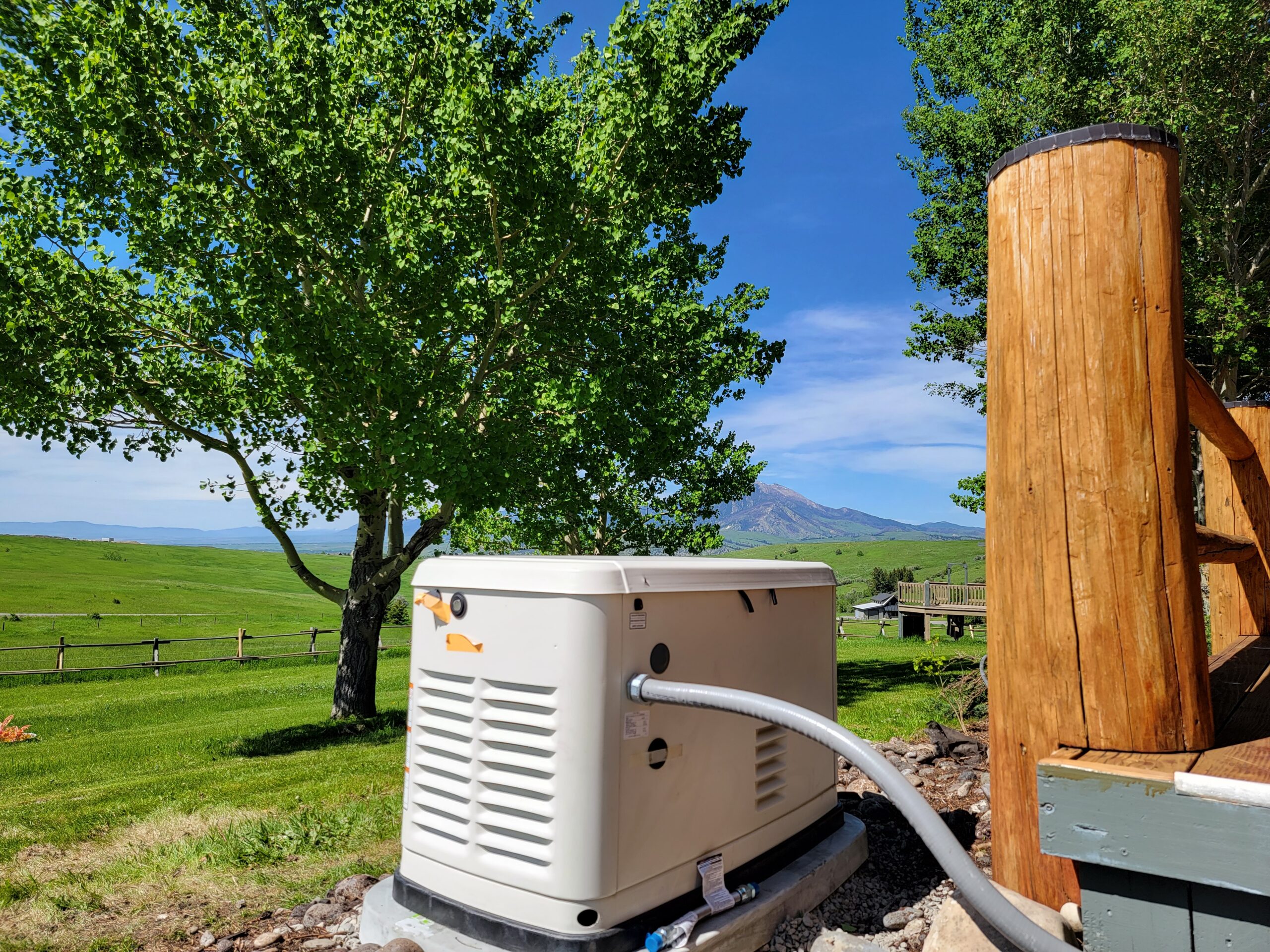 A standby generator, installed by an expert electrician, sits on a concrete pad next to a wooden deck, with a tree and open grassy field in the background under a clear blue sky.